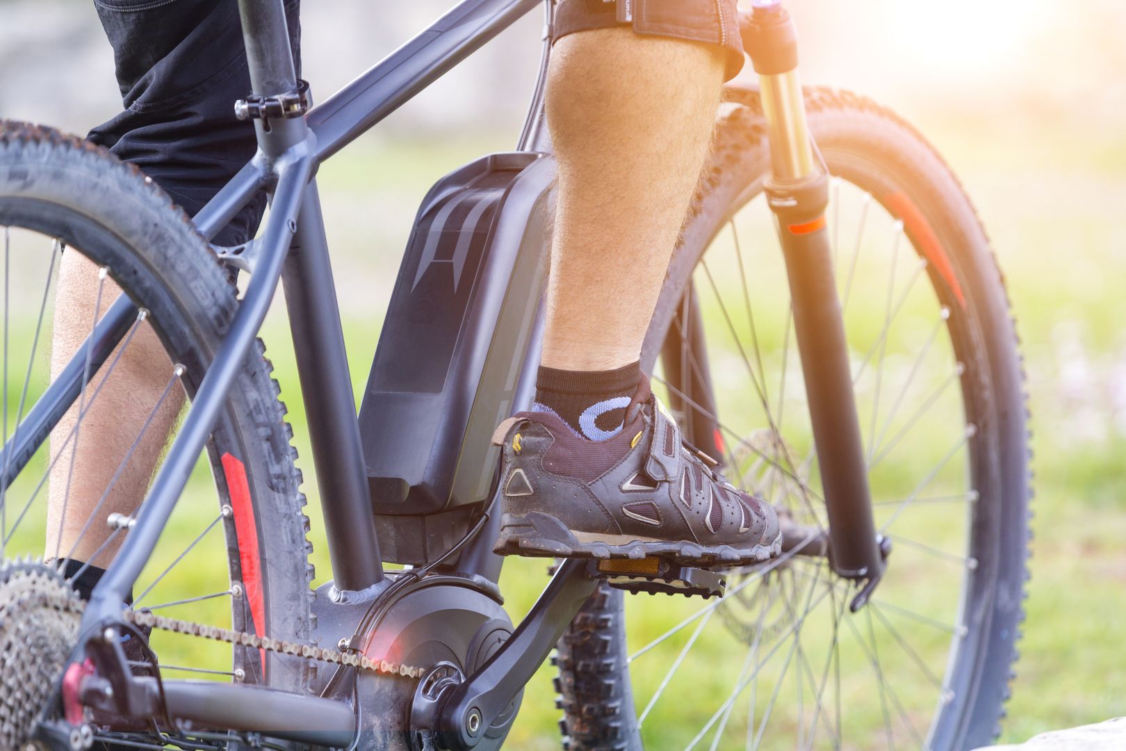 Nahaufnahme eines E-Bike-Fahrers mit schwarzem Fahrrad, Fokus auf Pedale, Motor und Akku bei sonnigem Wetter.